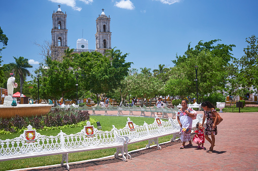 Two Mayan women and a little girl walk through the Parque Principal Francisco Cantón Rosado, the main square of the Mexican city of Valladolid, in the state of Yucatan. The church of San Servacio is in the background.