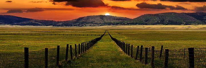 A fence lined panoramic view is inviting the viewer to walk in the grassy field toward the sunrise peaking above the mountain range in New Mexico.