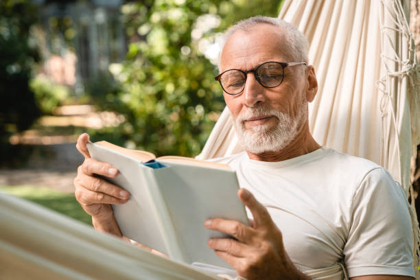 uomo concentrato del nonno che si rilassa riposando in amaca mentre legge il libro - men reading outdoors book foto e immagini stock