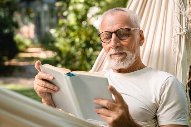concentrated grandfather man relaxing resting in hammock while reading book - lido imagens e fotografias de stock