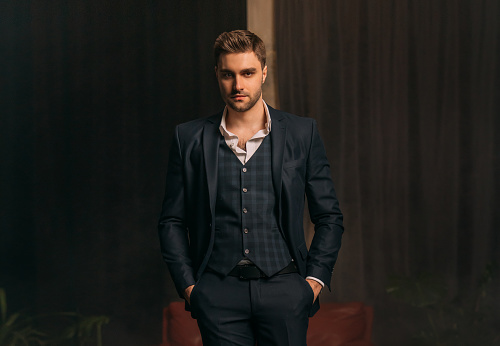 Portrait Handsome brutal man in classic strict three-piece suit jacket, trousers white shirt dark blue checkered vest. Successful businessman sexy guy stubble on face looks at camera. dark black room.
