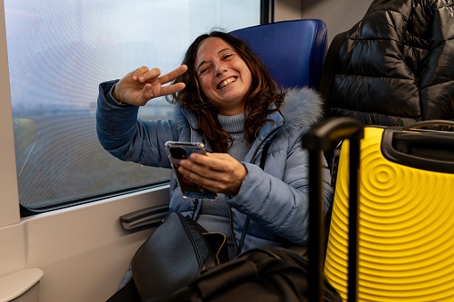 pretty middle-aged woman traveling on the train sitting among the luggage next to the window using the smart phone - vacation and fun concept