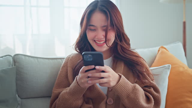 Happy young asian woman surfing social media on mobile phone, enjoying amazing news , Female having fun playing games chatting in social media sit on couch at home