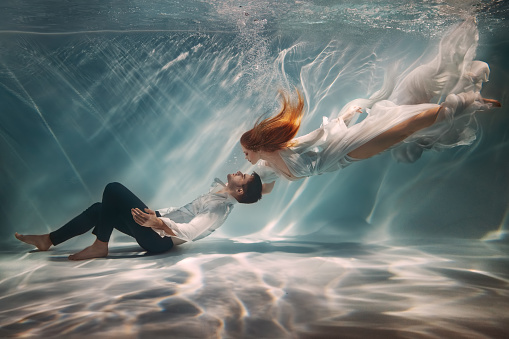 Real People art photo. Happy couple in love swim underwater, woman muse inspires male writer poet creator. Nymph girl saves drowning guy at bottom sea under water. Red hair white long silk dress float