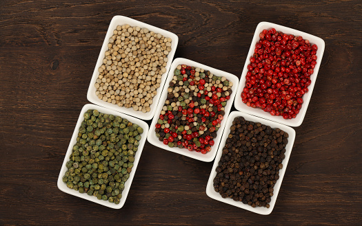 White ceramic bowls of assorted peppercorns, black, green, pink, white and mixed over dark wooden table background with copy space, elevated top view, directly above