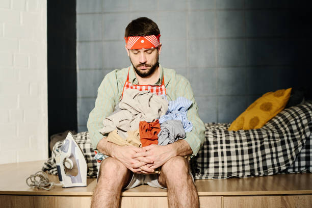 Tired young man with pile of crumpled clothes in hands sitting on edge of his bed in front of camera and looking at them