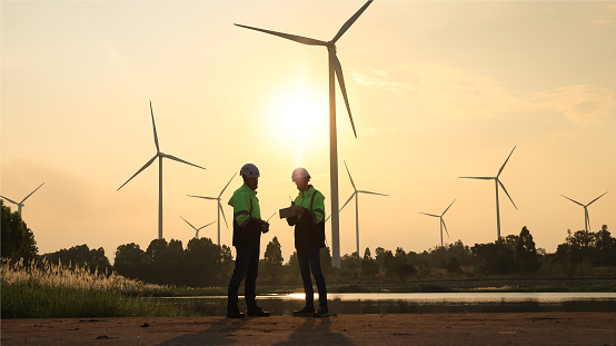 In silhouette two engineers planning maintenance on wind turbine equipment monitoring through digital device at windmill turbine farm