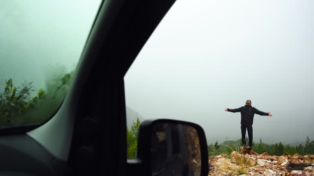 The man visible through the car window is smiling and enjoying the rain, opening his arms.