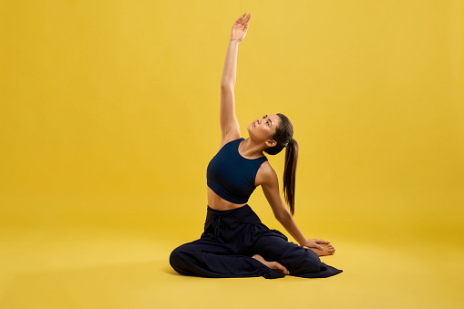 Front view of flexible, pretty woman sitting in pose of yoga in studio. Slim girl in black activewear posing with arm, stretched in air, isolated on yellow studio background. Concept of yoga.