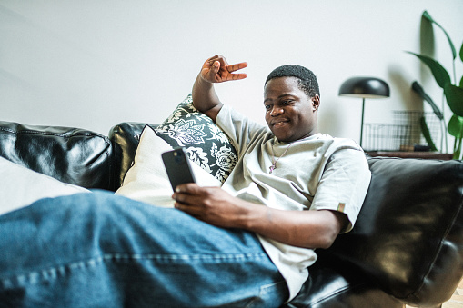 Young black man relaxing in living room sitting in cozy armchair and using smart phone