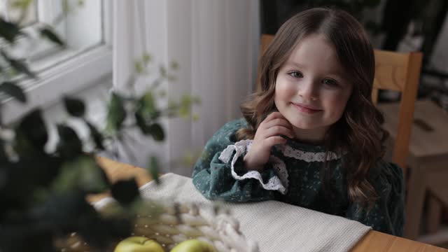 Adorable little girl with curly hair in rural style clothes sits by the table with green apples in woven tray, supports her chin with hand at home kitchen and looks at window. Happy childhood concept.