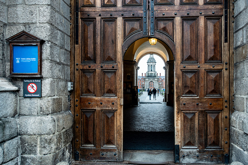 The image captures the open, ornate wooden doors of Trinity College in Dublin, Ireland, leading through a stone archway to reveal a glimpse of the iconic Campanile in the distance.