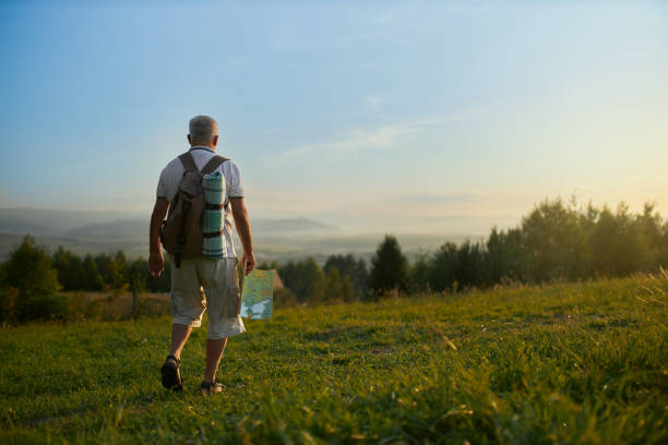 Middle aged man walking down hill, while hiking in mountains. Back view of adult male travelling with backpack and map along grassy hill, with scenic landscape on background. Concept of backpacking.