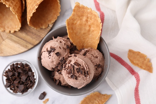 Bowl of tasty chocolate ice cream with chocolate chunks and pieces of waffle cone on white table, flat lay
