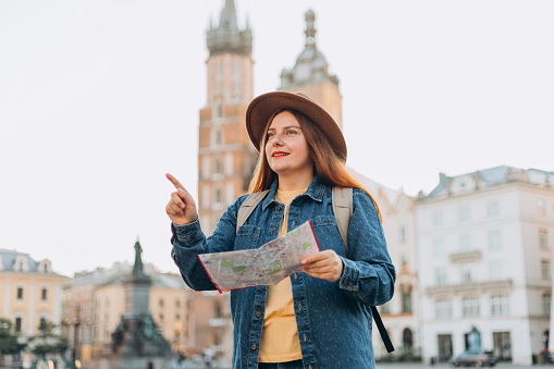 Attractive female tourist is exploring new city. Redhead woman pointing finger and holding a paper map on Market Square in Krakow. Traveling Europe in autumn. St. Marys Basilica
