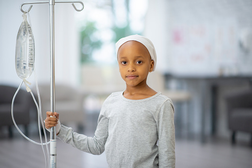 A young girl of African decent stands in her hospital room holding her IV pole as she receives her Chemotherapy treatment.  She is dressed casually, has a headscarf on to keep her warm and a neutral expression on her face.