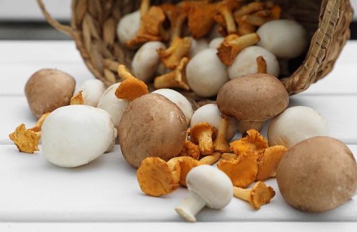Overturned wicker basket with different fresh mushrooms on white wooden table, closeup