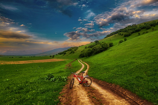 A bicycle is standing on a dirt road in the mountains against the backdrop of a beautiful evening sky, in a vertical frame.