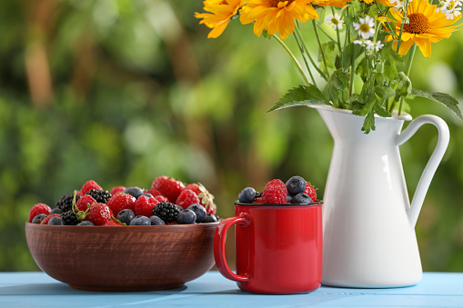 Different fresh ripe berries and beautiful flowers on light blue table outdoors