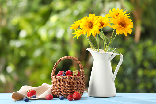 Wicker basket with different fresh ripe berries and beautiful flowers on light blue table outdoors