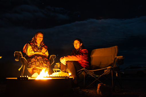 Under the dark sky at Furnace Creek campsite in Death Valley National Park, two women find solace and warmth by a crackling fire. A serene scene of friendship, laughter, and the quiet beauty of the desert night