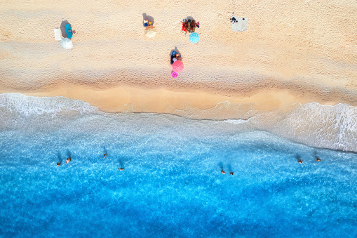 Aerial view of blue sea, waves, sandy beach and umbrellas with swimming people at sunset in summer. Tropical landscape with clear turquoise water. Top view from drone. Lefkada island, Greece. Travel