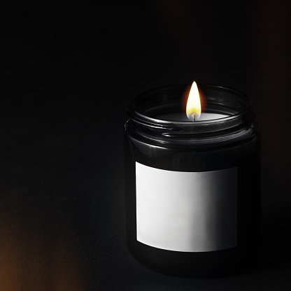 A lit decorative paraffin candle in a black glass jar on a black background. There is a blank white label on the can.