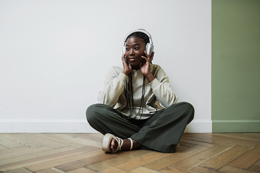 Beautiful black young woman enjoys listening to music standing near white and olive wall