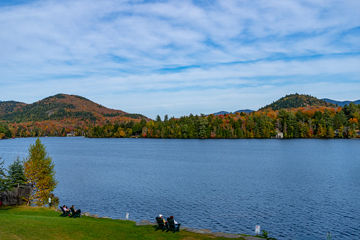 Autumnal leaf coloured forests near Mirror Lake, Lake Placid, New York State, USA.  There are people sat in wooden seats looking out over the lake.