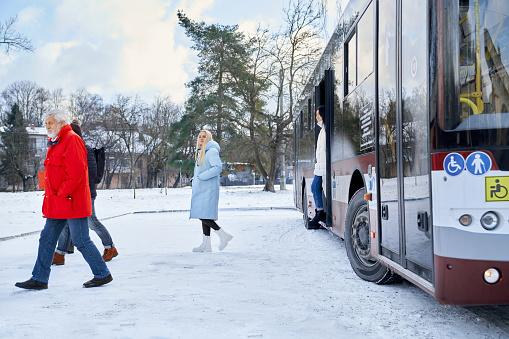Side view of elderly man in red jacket and blonde woman in blue wear leaving stopping bus in winter city. Full length of passengers getting off vehicle. Concept of transportation and city life.