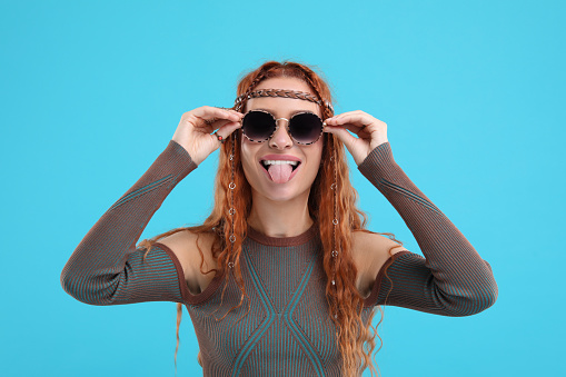 Stylish young hippie woman in sunglasses showing her tongue on light blue background