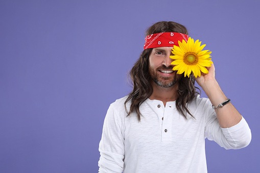 Stylish hippie man covering eye with sunflower on violet background, space for text