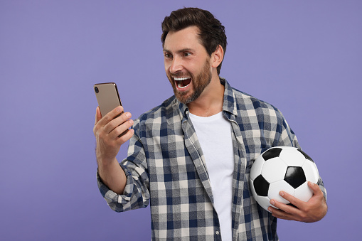 Emotional sports fan with soccer ball and smartphone on purple background