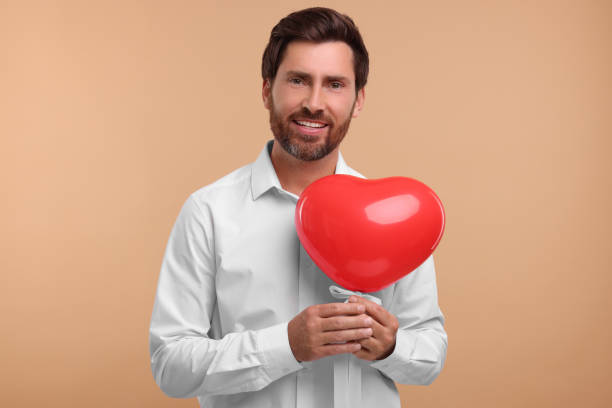 istock Handsome bearded man holding red heart shaped balloon on beige background 1935705449