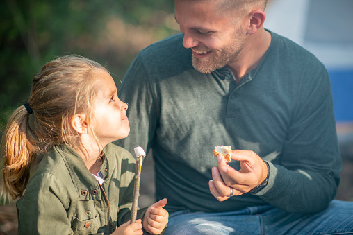 A sweet little girl sits beside her Dad as they roast Marshmallows around the camp fire.  They are both dressed casually and are enjoying the roasted treat.