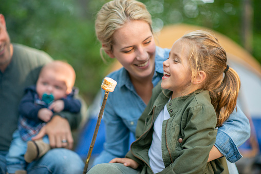 A sweet little girl sits up on her Mothers lap as they roast Marshmallows around the camp fire.  They are both dressed casually and laughing together.