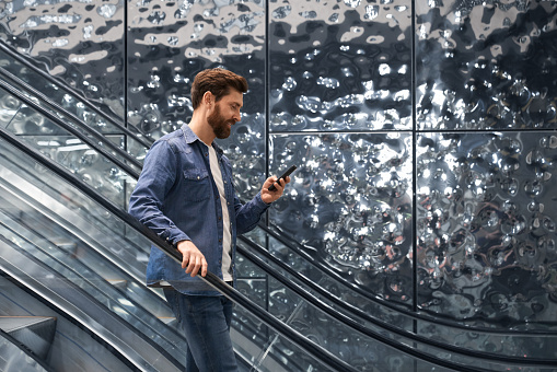 Smiling handsome man using smartphone, while going down on escalator of shopping mall. Side view of bearded male in denim jacket standing on moving staircase, while messaging. Concept of lifestyle.