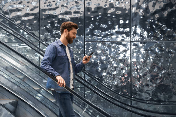 busy, smiling handsome man using mobile phone, while going down on escalator of shopping mall. - escalator shopping mall shopping transparent imagens e fotografias de stock