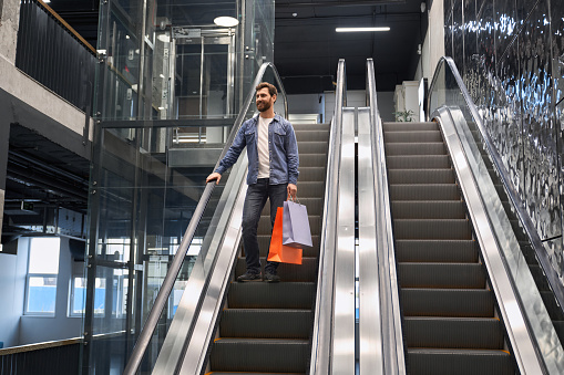 Smiling bearded man carrying shopping bags, while going down on escalator of shopping mall. Low angle view of happy male customer in casual clothes standing on moving staircase. Concept of shopping.