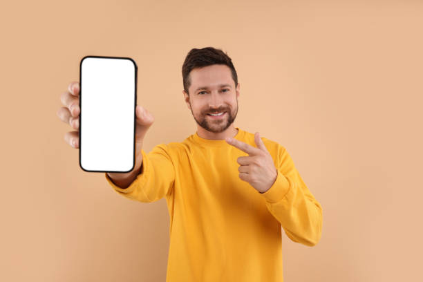 istock Handsome man showing smartphone in hand and pointing at it on light brown background 1935744936