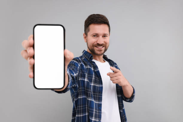 istock Handsome man showing smartphone in hand and pointing at it on light grey background 1935744954