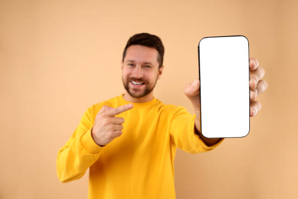 istock Handsome man showing smartphone in hand and pointing at it on light brown background, selective focus. Mockup for design 1935747051