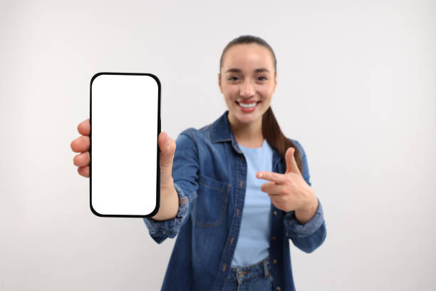 istock Young woman showing smartphone in hand and pointing at it on white background, selective focus. Mockup for design 1935748103