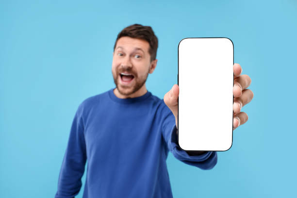 istock Surprised man showing smartphone in hand on light blue background, selective focus. Mockup for design 1935748116