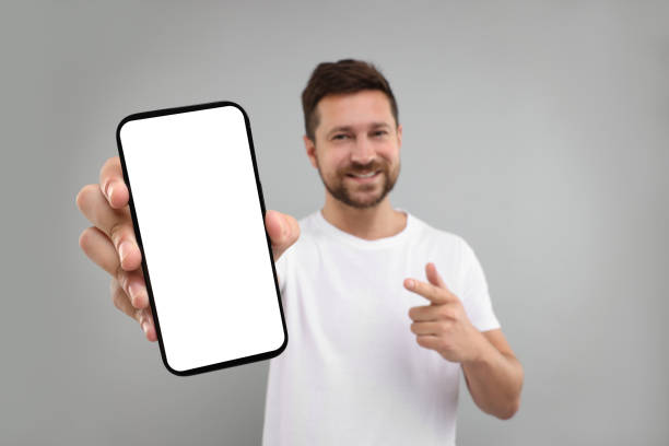 istock Handsome man showing smartphone in hand and pointing at it on light grey background, selective focus. Mockup for design 1935748246
