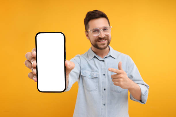 istock Handsome man showing smartphone in hand and pointing at it on yellow background, selective focus. Mockup for design 1935748252