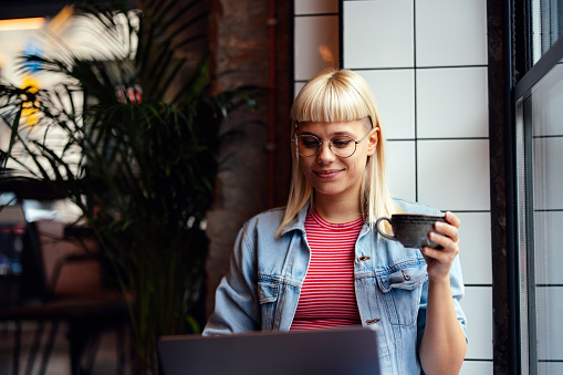 Portrait of a beautiful young woman in her 20s, sitting by the window in the cafeteria, working on her laptop computer and holding a cup of coffee. She is looking down at the screen and smiling.