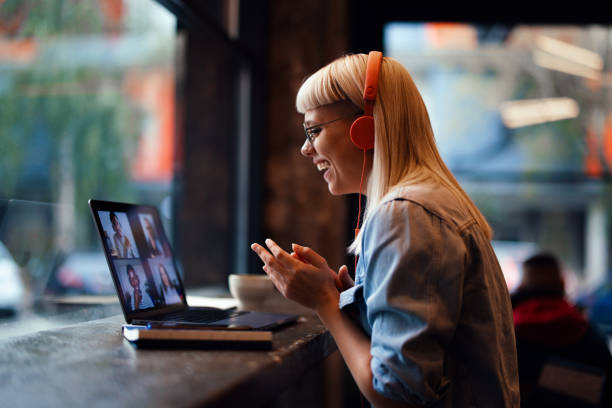 Portrait of a Cheerful Beautiful Young Woman Having an Online Meeting on Her Laptop in the Cafeteria (Copy Space) Close up shot of a beautiful young woman wearing glasses and headphone is sitting in the cafeteria. She is having a video meeting and is smiling while looking down at the screen. remote location stock pictures, royalty-free photos & images