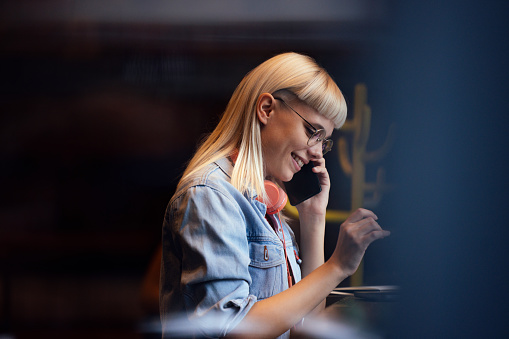 Close up shot of a beautiful young woman with blond hair and glasses, wearing headphones around her neck, talking on her mobile phone and smiling. She is sitting in a busy public space or a cafe.