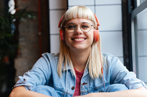Close up shot of a beautiful young woman with blonde hair, wearing glasses and headphones. She is looking at the camera and smiling.